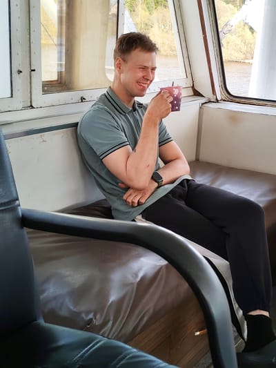 A man is seated on a dark brown bench within a cabin, possibly on a boat. He is wearing a short-sleeved gray polo shirt with a black trim, black sweatpants, and dark socks. On his left wrist, he has a watch. He is holding a polka-dotted cup, and there's a spoon sticking out of it. He is smiling slightly and looking toward the cup. The bench is located in front of a white wall and a window. The scene visible through the window appears to be a body of water with land and trees in the background. The lighting is natural. The location is identified as being in the city of Фёдоровка, Russia. A black chair is visible in the foreground.