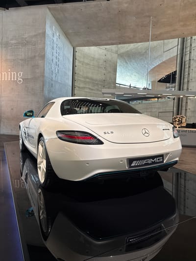 This media file shows the rear of a white Mercedes-Benz SLS AMG parked indoors. The car is positioned on a reflective surface, creating a mirrored image. The setting appears to be a museum or exhibition space with concrete walls and ceiling. The car's design features are prominent, including the "AMG" branding on the rear, the distinctive taillights, and the circular Mercedes-Benz logo. The rear grille has a mesh pattern. Next to the car, a section of a gray car is visible.
In the background, there are elements of industrial architecture and what looks like another vehicle engine on display. There are also vertical lighting fixtures and some signage. On a concrete pillar to the left of the car, there is a sequence of numbers engraved: "1 2 3 4 5 6 7 8 9 0" in one column and "2 3 4 6 7 10" in another. Another word, "ning" is visible below this. Above the car, a sign with German text is partially visible, including "und Lösung" and "tution". The overall impression is one of a well-lit, modern exhibition space showcasing automotive design and engineering.