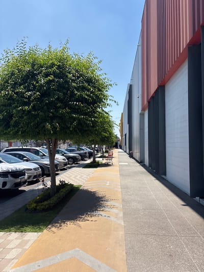 A street in San Pedro Cholula, Mexico, features a building with a red and white striped overhang. Adjacent to the building is a gray concrete sidewalk. The sidewalk is adjacent to a street of parked cars that have small trees on the left side of the cars.