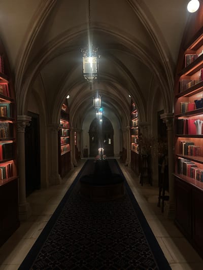 The scene depicts a long, dimly lit, gothic-style library hallway in Cernay-la-Ville, France. A vaulted stone ceiling spans the corridor, supported by stone pilasters. Light-toned stone tiles cover the floor, centrally traversed by a dark blue runner rug featuring a repetitive geometric pattern. Built-in wooden bookshelves, illuminated by integrated warm strip lighting, line both walls, displaying numerous antique-bound books of various sizes and colors. Multiple ornate, dark metal lanterns with glass panels hang sequentially from the ceiling, providing primary illumination. Approximately midway down the visible hallway, a circular wooden ottoman with a dark cushion is centrally positioned on the rug, supporting a candelabra with three lit candles. At the far end, a substantial, dark, carved wooden door is visible. No persons or actions are present within the scene.