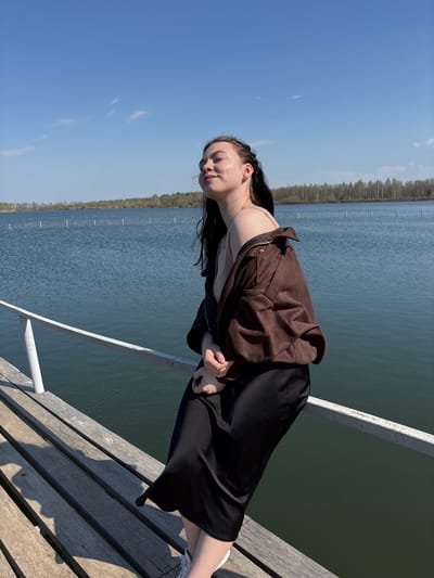 A young woman stands on a wooden pier near the water's edge. She wears a brown jacket over a light-colored top and a black skirt. The pier has a white railing on the left. Behind her, a large body of water stretches to the horizon, where trees line the opposite shore. The sky is blue with few clouds. The location is Crokhovo, Russia.