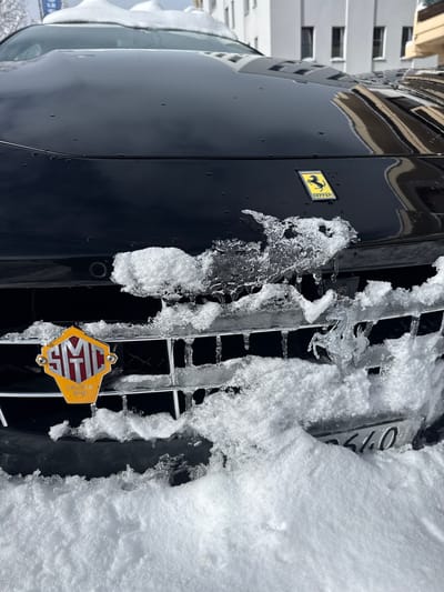 A black Ferrari automobile is prominently featured in a low-angle front view, parked outdoors in Sankt Moritz, Switzerland. The vehicle's front hood, grille, and bumper are heavily encrusted with snow and various ice formations, including dripping icicles. A yellow Ferrari 'prancing horse' emblem is centered on the black hood. Below this, an octagonal yellow and red 'SMC CRESTA RUN' badge is affixed to the left side of the front grille. To the right, an elaborate, clear ice sculpture replicating the Ferrari 'prancing horse' is present on the grille. Front parking sensors are visible, partially obscured by ice. Snow covers the lower grille and bumper, partially revealing the license plate with visible digits '2640'. The glossy hood exhibits water droplets, suggesting a thawing process. In the background, contemporary multi-story buildings with reflective windows stand under a bright daylight sky, characteristic of a winter resort environment.