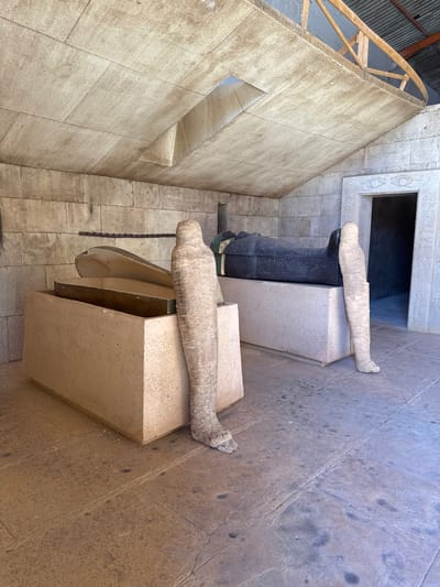 This image displays an ancient Egyptian tomb set, likely a film studio exhibit, located in Ouarzazate, Morocco. Two funerary setups, each comprising a sarcophagus on an elevated rectangular pedestal, occupy the foreground. On the left, a metallic gold/bronze sarcophagus rests on a light beige pedestal, its lid partially open to reveal a dark interior. A tan, textured mummy-like figure stands upright against this pedestal. On the right, a closed, dark grey/black sarcophagus with gold and green pharaonic accents sits on a light grey pedestal. A lighter beige, textured mummy-like figure stands upright against this pedestal. The environment features faux stone block walls and a flat, light-colored paneled ceiling with a central rectangular opening. An exposed wooden truss structure is visible above the ceiling panels in the upper right, indicating set construction. The floor is paved with worn, light grey rectangular tiles. A recessed doorway on the right wall is decorated with hieroglyphics and an "Eye of Horus" symbol. No human figures are present.