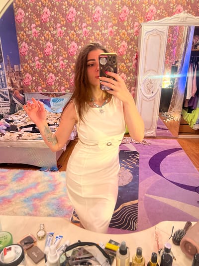 The image depicts a young woman taking a selfie in a bedroom. She is wearing a white sleeveless dress and a necklace with a snowflake pendant. Her hair is brown and shoulder-length. She is holding a smartphone in her right hand, using it to capture the reflection in a mirror. The room is decorated with floral wallpaper, a bed with a patterned duvet, a fluffy rug with pastel colors, and a large white wardrobe. Various items are visible on a table in the foreground, including a watch, cosmetics, and brushes. The overall impression is of a personal, somewhat cluttered, and vibrantly decorated living space. The context provided by Krasnoe-na-Volge, Russia, suggests a domestic setting within that geographical location.