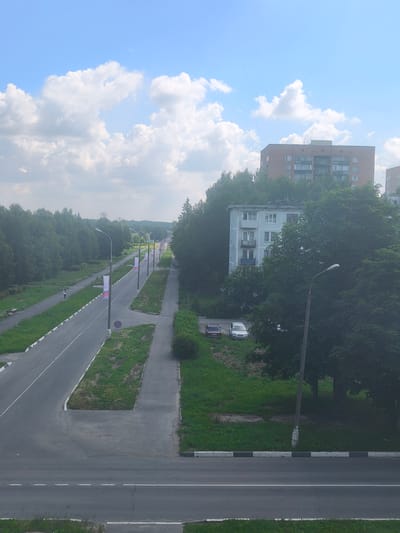 The image shows an elevated view of a street in Chekhov-2, Russia, under a partially cloudy sky. A paved road with white lane markings runs diagonally from the foreground towards the background, bordered by grassy areas and sidewalks. Several lampposts line the right side of the road, each adorned with a Russian flag. In the mid-ground, two cars, a red sedan and a silver hatchback, are parked on a grassy slope. Behind the road, a light blue, three-story apartment building is partially visible through dense green trees. Further in the background, a taller, brick apartment building with multiple balconies is visible, along with more trees and a hazy, distant landscape. The scene appears to be a residential area within the town.