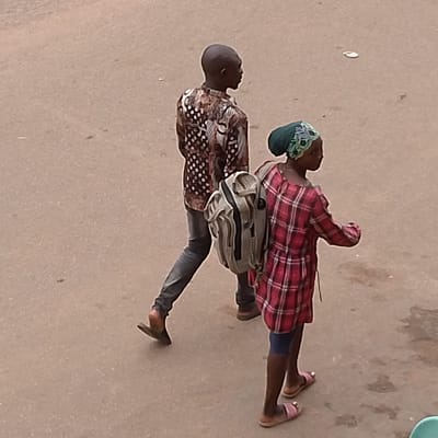 Two individuals, a male and a female, are walking on a paved surface, possibly a street or sidewalk, in Kayseri, Turkey. The male, on the left, is wearing a patterned shirt and dark pants, carrying a backpack. The female, on the right, is wearing a red plaid dress and a headscarf, also with a backpack. They appear to be walking in the same direction. The environment is a neutral, outdoor setting with a textured, light-colored ground.