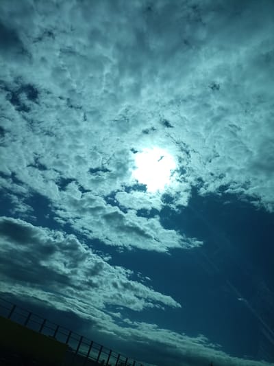 The image captures a dynamically colored sky above an unspecified elevated structure in Průhonice, Czechia. The sky is dominated by a deep teal and cyan hue, suggesting either specific lighting conditions or post-processing. A bright, overexposed sun is visible in the upper-left quadrant, shining through a significant break in the clouds. Sunlight diffuses through the atmosphere, with faint sunbeams visible to the right of the primary light source. The sky is largely covered by a multitude of altocumulus or stratocumulus clouds, varying in luminosity from bright white where illuminated by the sun, to various shades of grey and dark teal where thicker or in shadow. Patches of darker, clearer blue sky are discernible between the cloud formations. In the bottom-left corner, a segment of a yellow structure, possibly a wall or building facade, is present. Extending diagonally across the lower part of the frame, from the bottom-left to the mid-right, is a dark metal railing, consistent with that of a bridge, overpass, or elevated walkway. No individuals or specific actions are visible within the frame.