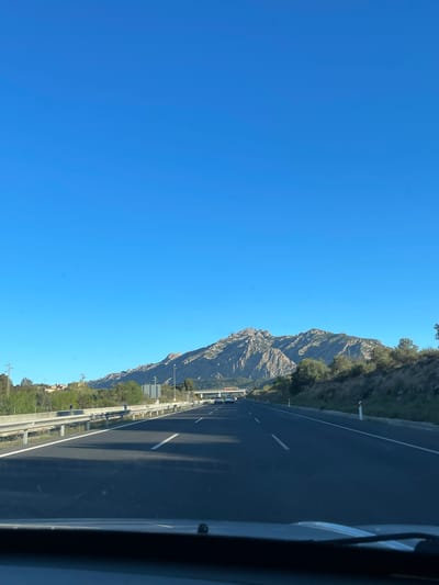 The image captures a view from a vehicle moving on a highway near Esparreguera, Spain. The highway is adjacent to a mountainous region. The sky is clear and blue. The highway is dark asphalt with white lane markings. A metal guardrail separates the road from the mountainside. The mountains are rocky with sparse vegetation.