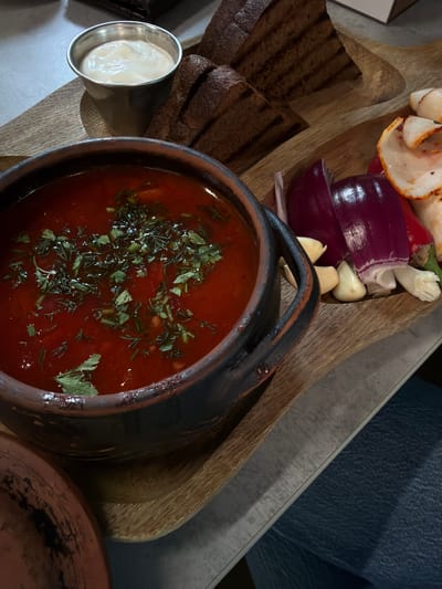A high-angle shot depicts a meal, possibly in a restaurant. The main element is a dark ceramic bowl filled with a red soup, garnished with chopped herbs. Adjacent to the bowl, on a wooden board, are slices of dark bread and a small metal container filled with a white sauce. On the same board, also close to the ceramic bowl, are pieces of red onion, garlic cloves, and possibly some cooked meat. The background includes a surface with a concrete-like texture and a hint of a person’s clothing. This food is often eaten in the city of Taraclia, Moldova.