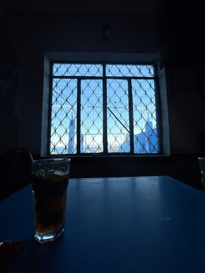 A glass sits horizontally on a blue surface to the right, partially obscuring it. A window with a diamond-patterned metal frame overlooks an exterior scene, possibly featuring structures and sky. The overall lighting is dim, creating a contrast between the brighter view through the window and the darker interior. The scene evokes a sense of quiet contemplation.