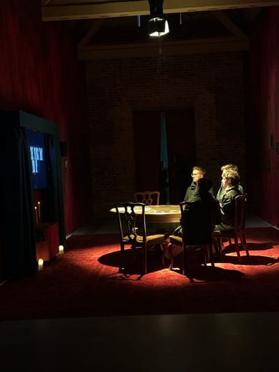 The scene depicts an interior room located in the City of Westminster, United Kingdom, characterized by a dominant dark red aesthetic. The walls are covered in textured dark red fabric, with a section of exposed red brickwork forming the rear wall. The floor is covered by a patterned dark red carpet in the illuminated area, with a darker, possibly wooden, floor in the immediate foreground.
An overhead spotlight casts a bright circle of light onto a central, ornate wooden table and the surrounding area. Four individuals are seated at this table, partially illuminated. One person, wearing glasses, is positioned facing left from the viewer's perspective. To their right, another individual with lighter hair is seen in profile. A third person is seated with their back partially turned towards the viewer, wearing dark attire. A fourth individual is largely obscured. The chairs are wooden with decorative backs.
On the left side of the room, a structure resembling a booth or cabinet, draped with dark green curtains, displays a brightly lit screen. The screen exhibits abstract light-colored shapes or patterns on a dark background. Below this screen, on a small wooden stand, two lit white taper candles provide additional soft illumination. Two further lit white votive candles are placed on the carpet directly below.
In the background, a dark arched doorway reveals a glimpse of a solitary figure standing in silhouette, partially obscured by darkness and a sliver of distant light. The overall lighting is dramatic and low-key, emphasizing the central gathering.