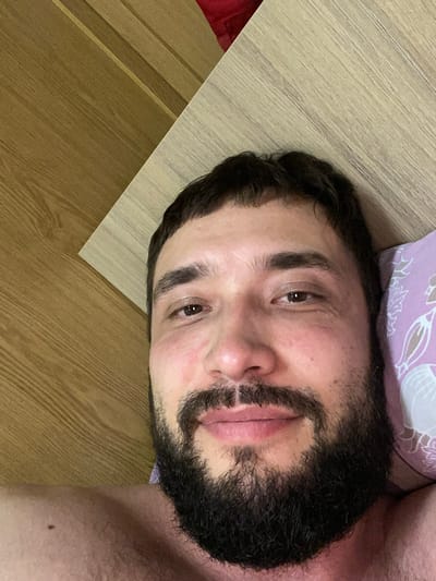 A close-up, overhead selfie of a man lying down. He has dark hair, a thick beard and mustache, and is looking towards the camera. He is lying on a wooden surface with a pillow visible on the right side of the frame. The pillow has a floral pattern in light purple and white. The man's bare shoulders are visible at the bottom of the frame. There is a mole on his left shoulder. The background consists of a wooden surface that appears to be a headboard or wall, with a distinct wood grain pattern. The lighting is soft and natural. The setting appears to be indoors, likely a bedroom.