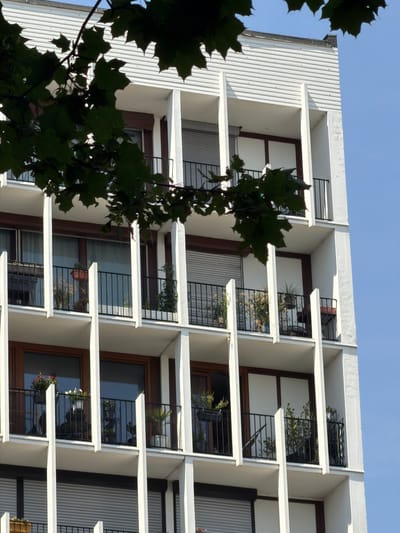 This image captures a section of a modern apartment building with a striking, geometric design. The building features a white, textured facade with numerous balconies that jut out from the structure. These balconies are enclosed by black metal railings, and some contain potted plants, adding touches of greenery to the stark architecture. The balconies are arranged in a repeating pattern, creating a sense of rhythm and order.

The overall impression is one of clean lines and contemporary aesthetics. The building is seen against a clear blue sky, suggesting it is a sunny day. The lighting indicates it is likely daytime, possibly late morning or early afternoon, given the angle of the shadows.

In the foreground, dark green leaves from a tree partially obscure the view of the building, suggesting the presence of nearby vegetation and a potentially urban setting with some green spaces. The leaves are out of focus, acting as a natural frame for the architectural subject.

There is no visible text in the image, and no people or specific activities are depicted. The focus is solely on the architectural details of the building and its integration with the natural elements. The location context of Courbevoie, France, suggests this is likely a residential building in a suburban or urban area of France. The overall mood is serene and architectural, highlighting the interplay of form, light, and shadow.