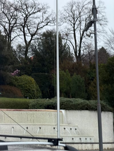 This image forensically depicts an outdoor urban park scene on an overcast day in Ouchy, Lausanne, Switzerland, specifically featuring the Fontaine de la Navigation (also known as Fontaine des Pêcheurs) located on Quai d'Ouchy.

In the immediate foreground, the top section of a vehicle, including dark roof rails, is partially visible. A silver metallic flagpole stands vertically on the left side of the frame, extending upwards. On the right, a dark grey utility pole is present, fitted with an angular, grey-colored device, possibly a security camera or sensor, pointing towards the left.

The central subject is a multi-tiered fountain, constructed from light-colored, vertically grooved concrete or stone. The fountain's upper and middle tiers feature multiple spouts actively emitting horizontal streams of water into collecting basins below. Behind the fountain, a steep, landscaped embankment rises, densely covered with a variety of dark green evergreen shrubs and lighter green ground cover.

The background is dominated by numerous large, bare deciduous trees, indicating a late autumn or winter season. A small cluster of reddish-pink flowering foliage is visible near the left edge of the embankment. A structure with a reddish-brown roof is faintly discernible through the trees in the distant background. The sky is uniformly overcast and grey. No human figures or overt interactions are visible within the frame; the primary action is the flowing water from the fountain.