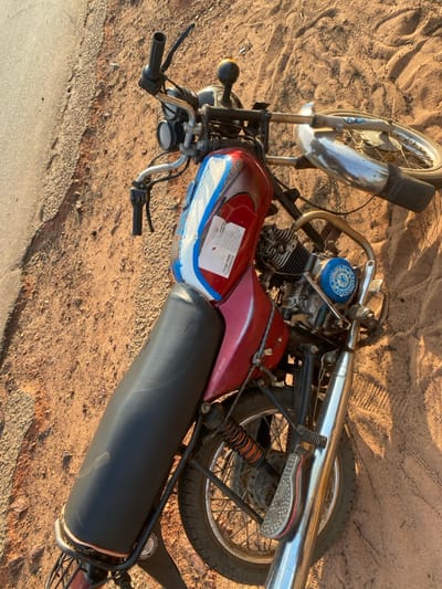 A red and blue motorcycle lies on its side in the sand. The black seat is prominent, and the engine has a blue top. The front wheel faces upward, while the rear wheel is tilted towards the ground. Chrome exhaust pipes are visible. A sticker on the fuel tank displays information about governance.