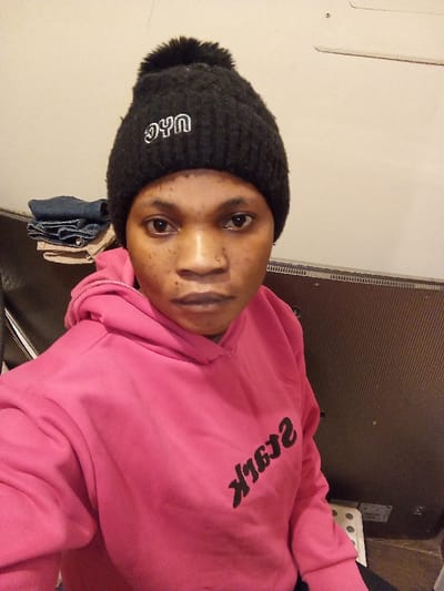 An individual is depicted indoors in Madinaty, Egypt. The subject, a person with dark skin, dark eyes, and a small metallic stud in the right nostril, is positioned facing directly towards the camera. They wear a black knit beanie topped with a pom-pom, featuring "NYC" embroidered in white letters on the front. Below the beanie, a bright pink hooded sweatshirt is worn, displaying inverted black text that reads "STARk" across the chest. The background on the left includes a stack of folded dark blue fabric situated on a surface, partially obscured by a dark, possibly upholstered, seat or chair back. The background on the right features a plain, light-colored wall above a dark, vertically ribbed or textured panel. A small, light-colored rectangular object with several dark circular marks is partially visible at the bottom right corner of the frame, below the dark panel.