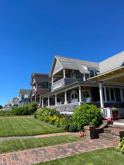 This is a photograph taken on a sunny day in Oak Bluffs, Massachusetts, United States of America. The image displays a row of colorful Victorian-style houses with intricate architectural details, characteristic of the Martha's Vineyard gingerbread cottages. The houses are situated along a street, with manicured green lawns and vibrant flower beds in the foreground. The sky is a clear, deep blue. A brick pathway leads up to the porch of one of the houses, which features white rocking chairs and a yellow awning. The scene evokes a sense of charm and tranquility.