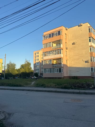 The image depicts a residential building in the city of Popovo, Bulgaria, bathed in the warm light of either sunrise or sunset, casting a long shadow across the building. The building, painted in shades of peach and cream, has multiple balconies with white railings and a series of windows. Visible on the building's exterior are at least three air conditioning units. In front of the building, a strip of overgrown grass runs along a paved walkway, separating it from a street. Several cars are parked in the grassy area. A single, yellow building is partially visible on the left side of the image. Overhead, a tangle of dark electrical wires crisscrosses the clear blue sky. The overall scene suggests a typical urban residential setting.