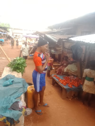 The image captures an outdoor market scene, likely in a rural or semi-urban setting, characterized by earthy ground and makeshift stalls. The ground is a reddish-brown dirt path.
In the foreground, slightly to the left of the center, a young person is standing in profile, facing right. They are wearing a black beanie, an orange short-sleeved t-shirt with blue and white striped accents on the sides, and blue athletic shorts with white stripes. On their feet, they wear blue flip-flops. They are holding a small, light-colored object to their mouth, possibly a lollipop or snack, and appear to be smiling or engaged. To their left, on a raised structure, is a large pile of fresh green leafy vegetables. Near their feet, a yellow plastic jerrycan and a blue tarp covering unknown items are visible.
To the right of the main subject, there is a market stall. A wooden table, covered with a blue tarp, displays a large quantity of red tomatoes for sale. Some other dark-colored produce is also visible on the table. Behind this stall, a person is seated, partially obscured, and further to the right, a small child in a light-colored dress with a green sash is standing. The market stalls in the background are constructed with corrugated metal sheets and wooden supports, indicating simple, utilitarian structures.
In the mid-ground and background on the left, several other people are walking along the dirt path. A man in a light-colored shirt and dark pants is visible walking towards the viewer. Distant figures and more market structures with various coverings can be seen further down the path, blending into the bright, almost washed-out background sky. A wooden pole leans diagonally in the lower-left foreground, part of the market's infrastructure.
The lighting is bright, suggesting daytime, possibly under an overcast sky or very strong sunlight leading to overexposure in the upper part of the image. The overall impression is that of a bustling local market focused on fresh produce. No discernible text is visible in the image.