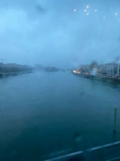 A blurry, rainy window view showcases a cityscape along a body of water. Tall buildings line the distant shore, partially obscured by fog. Traffic moves along the waterfront. Droplets distort the scene on the glass. The sky is overcast and dark. A linear structure is visible in the foreground, possibly part of the window frame.