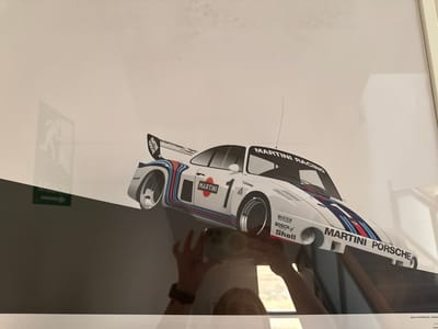 The image is a close-up shot of a framed print featuring a white Martini Racing Porsche race car with its iconic blue, red, and white livery. The car is depicted from a low, side-angled perspective, with its distinctive rear wing prominently displayed. The background is a plain white wall, with a subtle shadow of an exit sign visible on the left. The lower portion of the print transitions to a dark gray, angled surface.
Text visible on the print includes "MARTINI RACING," "MARTINI," "Turboleader," "DUNLOP," "BILSTEIN," "BOSCH," "Shell," and "MARTINI PORSCHE." There is also some text at the very bottom, partially obscured, but appearing to include "ROLF STOMMELEN | PORSCHE".
The photo appears to have been taken indoors, possibly in a home or office, given the framing and the reflection of a person holding a phone in the foreground, suggesting the photo was taken with a mobile device. The lighting is bright, casting reflections on the glass of the frame. No specific time of day or weather conditions are discernible, but the indoor setting implies these are not primary factors. The overall impression is of a display of automotive memorabilia. The location context provided is Getafe, Spain, but this cannot be visually confirmed from the image itself.