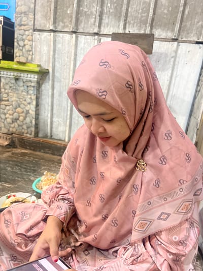 In the city of Wedarijaksa, Indonesia, a young Indonesian woman wearing a pink hijab and matching attire with an "SS" logo pattern is captured in a close-up portrait. She is seated, with her head tilted downwards, appearing to be engrossed in her mobile phone held in her hands. A small plate with food and a green bowl of what looks like crackers or snacks are visible to her left. The background features a rustic wall made of corrugated metal sheets and a textured stone wall. The lighting suggests it is either dusk or an indoor setting with artificial light.