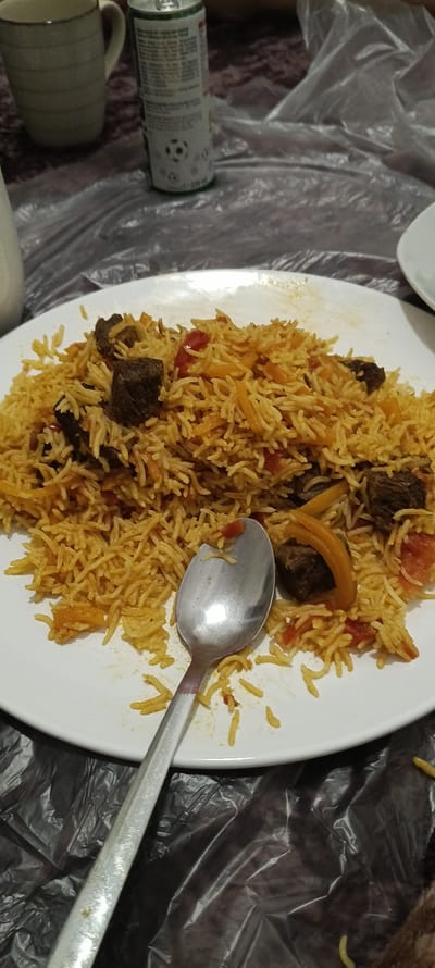The image shows a plate of food, likely a rice dish with meat, placed on a table covered with a transparent plastic sheet. A silver spoon rests on the food, suggesting that a meal is being consumed. In the background, a tall, ceramic mug and a partially visible soda can are present. The soda can has some text and a graphic of a soccer ball. The scene appears to be indoors, possibly in a casual dining setting. The lighting is subdued, and the focus is primarily on the food. There are no discernible people or specific architectural details visible that would indicate a precise location, apart from the provided context of Linz, Austria. The overall impression is that of a simple meal being enjoyed at a table.