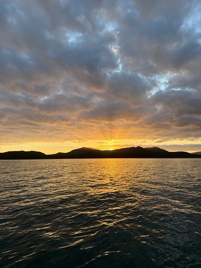 The image displays a wide-angle view of a sunset over a body of water and a distant landmass. The scene is located in a coastal area of Dumbéa, New Caledonia, France.
The upper portion of the frame is dominated by a sky filled with scattered altocumulus or stratocumulus clouds, which are illuminated with hues of orange and yellow near the horizon, transitioning to darker grey and muted blue tones higher up. Crepuscular rays emanate from a bright, partially obscured sun positioned just above the distant landmass, slightly right of the frame's horizontal center.
A dark, silhouetted mountain range or a series of hills forms a distinct horizon line, separating the sky from the water. The profiles of these landforms are irregular.
The lower portion of the image depicts a body of water, likely the ocean or a large bay. Its surface is textured with numerous ripples and small waves. The setting sun's light reflects intensely on the water, creating a broad, shimmering path of golden-orange light that extends from the horizon towards the foreground. The water appears darker in the immediate foreground and towards the left and right edges, receiving less direct light reflection. No human figures, vessels, or artificial structures are visible within the frame.