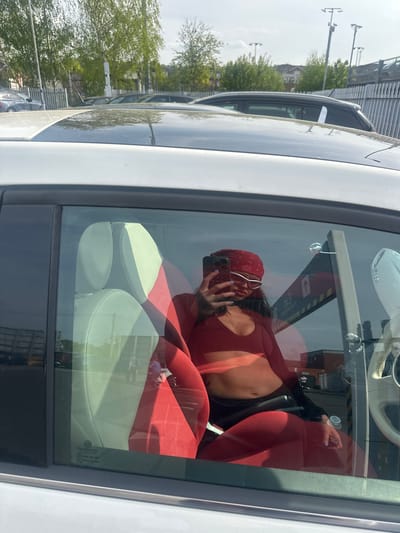 A woman wearing a red bandanna and red top is sitting inside a white car in what appears to be a parking lot. She is holding a phone and taking a picture of herself in the reflection of the car window. The car has red and white seats and a white steering wheel. Outside the car window, there are trees and utility poles in the background, along with buildings that suggest an urban environment in Sevenoaks, United Kingdom.