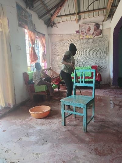 An indoor scene in Manda-Shivanga ward, Kenya, shows a room with a concrete floor exhibiting wet patches and stains. The ceiling features exposed wooden rafters and corrugated metal sheeting. Walls are light-colored, with a patterned white fabric covering a portion of the back wall. A window on the left has sheer orange/brown curtains. Two framed pictures adorn the left wall, and a poster depicting babies and a woman is on the right wall.
Two individuals are present:
1. A male subject is seated on a red-framed chair with green upholstery on the left, wearing a light-patterned shirt and dark trousers. His bare feet are visible.
2. A female subject stands center-right, wearing an olive-green t-shirt and dark trousers, with her head covered. She is holding a long stick or broom-like object, appearing to sweep or clean the floor. Her bare feet are visible, standing in a wet area.
In the foreground, a light blue wooden chair stands centrally, facing the viewer. An orange plastic basin containing liquid or debris rests on the floor to the left, near the window. The scene suggests a domestic environment undergoing cleaning, possibly in response to water accumulation on the floor.