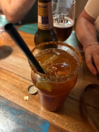 A close-up, eye-level shot focuses on a drink in a glass with a dark straw and a salted rim. The drink is a deep amber color, with ice cubes visible inside. A slice of lime is submerged in the liquid. The glass rests on a wooden table, which appears to be outdoors, possibly at a restaurant or bar in Begur, Spain. A beer bottle with a label that reads "FRED DAM TORIA" stands behind the glass. In the background, a person's arm is visible, along with another beer glass containing a dark liquid and foam. The overall setting suggests a casual dining or drinking experience.