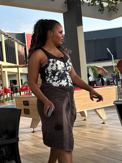 A woman stands outdoors on a tiled surface, likely a patio or poolside area, holding a smartphone up with her right hand as if taking a photograph or recording a video. She is partially wrapped in a brown towel and wears a black and white floral print swimsuit top. The background includes palm trees, umbrellas, chairs, tables, and multi-story buildings. Based on the prompt, the location is Rubuchi, Nigeria, although specific location is not identifiable from the image. Other people can be seen in the background.