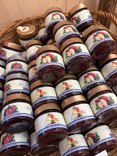 The image shows a close-up, eye-level view of a tightly packed wicker basket filled with numerous glass jars of jam. The jars have gold-colored lids and white labels with blue borders. The labels feature illustrations of fruits and berries, and text in Italian, indicating different jam flavors. Some visible flavors include "Frutti di Bosco" (forest fruits), "Mela e Ribes Nero" (apple and blackcurrant), and "Mela, More e Zenzero" (apple, blackberry, and ginger). The jars are stacked and angled, creating a dense composition. The background consists of the rough texture of the wicker basket and a hint of a wooden surface. The lighting is bright and slightly diffused, highlighting the contents of the jars and the details on the labels. No people or specific locations within Lateis / Latais, Italy are identifiable in the image.