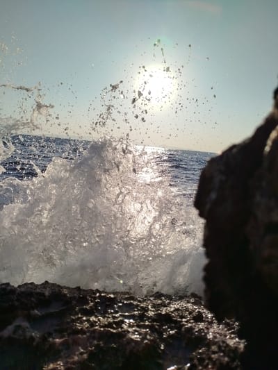 An extreme close-up captures a dynamic wave crashing against dark, wet rocks in Karaburun, Turkey. The sun, high in the sky, shines brightly, creating a lens flare and illuminating the spray of water droplets as they erupt from the impact. The ocean's surface is visible in the background, with a slightly hazy horizon. A large, dark rock formation dominates the right side of the frame, partially obscuring the view of the sea. The foreground is composed of rough, textured rocks, reflecting the sunlight. The image appears to have been taken during daylight hours, with the sun at its zenith. There are no people or animals visible in the scene.
