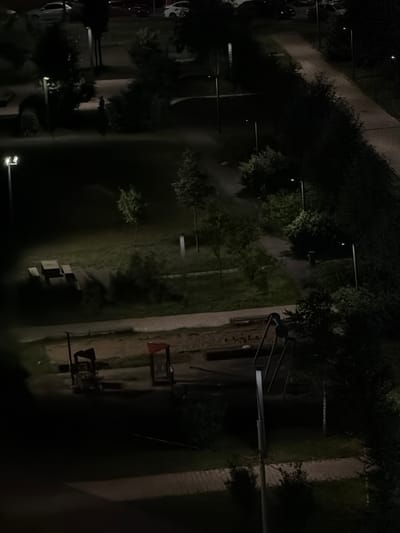 This image shows an aerial view of an outdoor area at night. In the foreground, there is a playground with red and blue structures and a sandpit. Further back, there are picnic tables and benches on a grassy area. Several trees and lampposts are visible throughout the scene. In the background, there is a paved area that appears to be a road or pathway, with cars parked along the side. The overall lighting is dim, with artificial light sources casting shadows. The location is identified as Bobrovo, Russia.