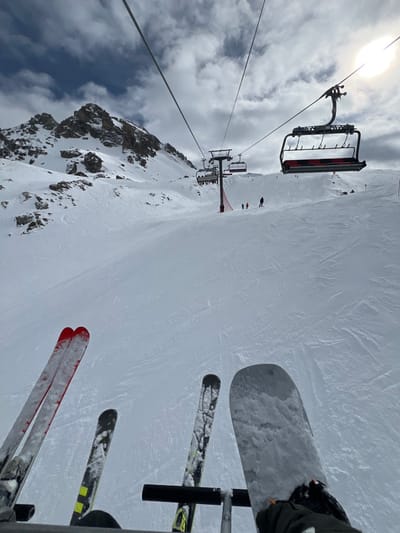 The original image, a rotated vertical shot, presents a first-person perspective from a ski lift ascending a snowy mountain, characteristic of a ski resort like Val-d'Isère, France.
In the immediate foreground, the skis of the person taking the photo are visible, secured to the footrest of the ski lift chair. One pair of skis is predominantly red, while another pair shows yellow and black accents. A snowboard is also prominently featured, attached to the same footrest.
Below and stretching into the distance, a wide, snow-covered ski slope is visible, marked with numerous faint tracks from previous skiers. Several small figures, identified as skiers or snowboarders, can be seen on the slope. Further down the mountain, another ski lift chair, occupied by multiple passengers, is seen approaching a red-accented support pylon.
To the left, a rugged, snow-dusted rocky mountain peak rises under a partly cloudy sky. The bright sun is visible in the upper left corner, creating a strong glare through the clouds. The scene captures an active daytime environment at a ski resort, with conditions appearing bright despite the cloud cover. There is no discernible text visible in the image.