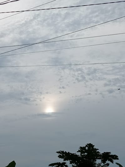 The image depicts an overcast sky with the sun partially visible through the clouds, casting a diffused light. Several dark power lines crisscross the sky horizontally. In the lower right corner, the silhouette of a leafy tree is visible against the sky. In the lower left, a portion of a large green leaf is also present. In the distance, a small dark shape, possibly a bird, is in flight. The scene is likely an outdoor environment in Kruengmanyang, Indonesia.