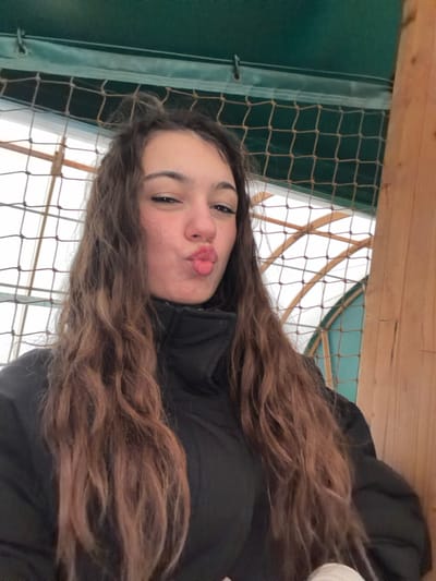 The image depicts a young female, approximately late teens to early twenties, posing for a close-up self-portrait in Souffelweyersheim, France. She has long, wavy, multi-toned brown hair with lighter streaks, falling over her shoulders and chest. Her eyes are open, and her eyebrows are dark and distinct. Her lips are pursed forward in a "kiss face" expression. She is wearing a black, high-collared puffer jacket. The background elements suggest an indoor or semi-enclosed facility. Directly behind her and above, a dark green structure or beam is visible, beneath which a dark-colored mesh net with a grid pattern is strung. Behind the net, translucent white or frosted panels form part of a roof or wall, supported by light-colored wooden beams that create an arched or angled structure. A vertical wooden pillar or beam is partially visible on the right side of the frame. The lighting appears to be ambient, likely from natural light filtering through the translucent panels.