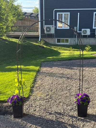 This is a photograph of a garden scene. In the foreground, two black square planters are placed on a gravel pathway, each filled with a dense cluster of purple flowers, possibly petunias, with a vine growing up a metal archway that stands between them. The archway is made of dark metal and has a curved top. To the left of the pathway, a well-maintained green lawn slopes upwards towards a wooden deck and fence. In the background, a dark blue house with white trim and multiple windows is visible. Two outdoor air conditioning units are mounted on the exterior wall of the house, along with a utility meter and conduit piping. The sky is partly cloudy, and sunlight casts shadows on the gravel and lawn. The scene appears to be a residential backyard, likely in Dieppe, Canada, given the architectural style and landscaping. There are no people or animals present in the image.