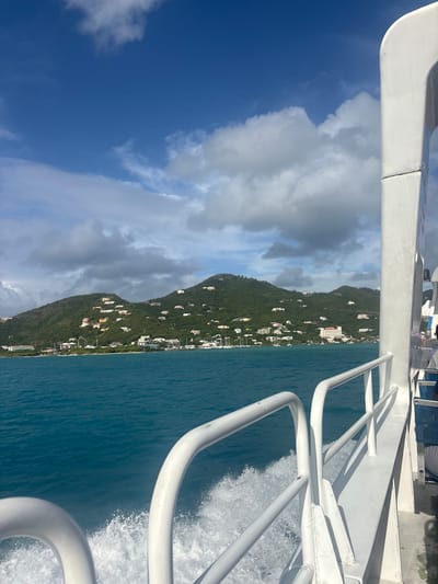 The scene depicts a view from a white vessel moving across turquoise water, oriented towards the city of Road Town, British Virgin Islands. In the immediate foreground, white boat railings and a section of the boat's hull are visible on the right, with a substantial white wake trailing from the vessel in the water below the railings. A partial view of seated individuals is discernible through a window or opening on the starboard side of the boat's interior. The midground features a wide expanse of clear, blue-green ocean. In the background, verdant, undulating hills, densely covered in tropical vegetation, rise from the coastline. Numerous white and light-colored residential structures are sparsely distributed across these hillsides, extending towards the crests. Along the immediate shoreline at the base of the hills, a concentration of buildings, including what appears to be a multi-story hotel or commercial complex, is visible. A bay or harbor area is present, dotted with multiple moored or anchored sailboats and yachts. The sky above is largely blue with scattered, fluffy white cumulus clouds, indicating fair weather conditions.