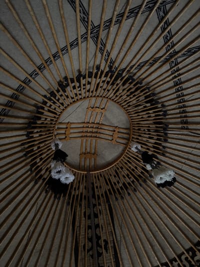 The image is an interior, worm's-eye view of a yurt, a traditional portable dwelling. The ceiling is constructed of wooden poles radiating from a central circular wooden frame, known as a "shanyrak." The poles are covered by a felt or fabric material, upon which a decorative band of black and white geometric patterns is visible. Several tassels, some white and some black, hang from the central frame. The lighting is dim, suggesting it might be nighttime or that the yurt is not well-lit. The setting is identified as Kyzyl-Korgon, Kyrgyzstan, a location known for its traditional nomadic culture and yurts. There are no people or animals visible in the image.
