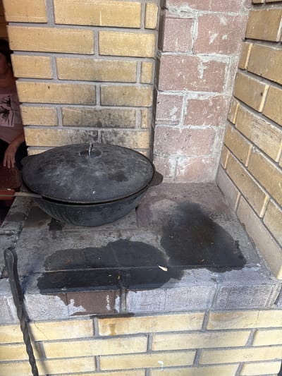 A large, black, cast-iron cauldron with a lid is placed in a brick oven. The cauldron is positioned to the left of the frame, with its lid slightly ajar, revealing a glimpse of its interior. The oven is constructed with yellow and pinkish-brown bricks, laid in a staggered pattern. The interior of the oven shows signs of use, with dark, sooty patches on the brickwork.

A metal handle, likely for the cauldron, protrudes from the lid, and a wooden-handled tool, possibly a knife, is placed on the edge of the oven. To the right of the cauldron, a dark, wet patch is visible on the floor of the oven, and a twisted metal poker rests on the brickwork. The background is filled with more brickwork, creating a textured and rustic setting.

The lighting suggests it might be daytime, but the specific time is not clear. The overall scene evokes a sense of outdoor cooking, possibly in a traditional setting, given the presence of the brick oven and cast-iron cauldron. A small portion of a person's arm and shoulder wearing a t-shirt can be seen in the lower left corner of the image. No visible text is present in the image. The location is contextually identified as Chernogorsk, Russia.
