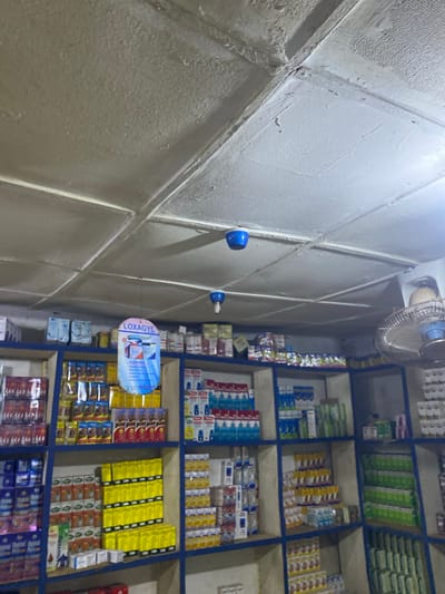 The media file shows a section of a small shop in Jere South, Nigeria, with shelves stocked with various products. The setting appears to be indoors, with a white textured ceiling and walls. On the ceiling, a ceiling fan is visible, along with two blue lights or sensors. Hanging from the ceiling by a wire is a blue and white oval sign that reads "LOXAGYL" and some other text that is difficult to discern.

The shelves are organized into compartments and filled with a variety of packaged goods, likely pharmaceuticals or consumer products. The packaging varies in color and design, with many items appearing to be in boxes. Some prominent colors include yellow, green, blue, and red. There are also some white and blue boxes stacked on the shelves.

The lighting suggests it might be daytime, but the indoor setting makes it difficult to determine the exact time. There are no people visible in the media file. The overall impression is of a neatly organized retail space in Nigeria.