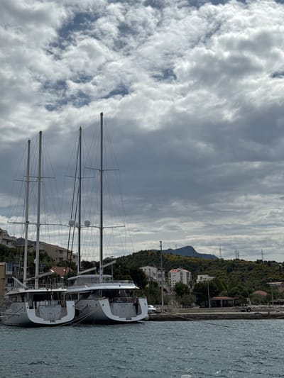 The image shows two white sailboats docked at a marina in Dugi Rat, Croatia, at the coordinates 43.446 latitude and 16.635 longitude. The boats are docked side-by-side with their masts pointing towards the cloudy sky. The boat on the left has the name "MANDILA" painted on the back, while the boat on the right has the name "OMNIA" painted on the back. The boats are docked in front of a low-lying shoreline with a paved area and a few small buildings. There are trees and bushes behind the buildings, and hills beyond that, covered in vegetation. The sky is overcast with many white clouds. The overall scene is serene and peaceful.
