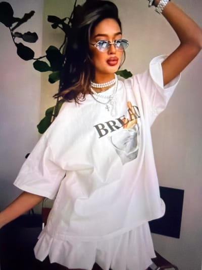 The image shows a young woman standing indoors, possibly in a residential setting in Beșalma, Moldova. She is wearing oversized white clothing, including a t-shirt with the word "BREAD" and a graphic of a loaf of bread in a pocket, and ruffled shorts. She also has on a pearl necklace and large, ornate sunglasses. Her left arm is raised, and she appears to be posing for the camera. A large leafy plant is visible in the background to her left. The overall lighting is soft and diffused.