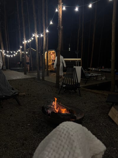 An outdoor scene at night in Ivachiv Horishnii, Ukraine, features a campfire in the foreground with logs burning. In the midground, a wooden deck is illuminated by string lights, with trees surrounding the area. A man in black shorts stands on the deck. Further back, a sauna structure with a rounded entrance is visible, and another man in a white robe stands inside. Two lounge chairs are present on the deck, one with a white towel draped over it. There is also a small table with drinks and what appear to be plastic bags. The ground is covered with gravel and pine needles.