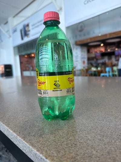 A green plastic bottle of Topo Chico mineral water, specifically the "Toronja" (grapefruit) flavor, lies horizontally on a light brown countertop with a slightly speckled texture. The bottle has a pink cap. The label is yellow and black, bearing the brand name and indicating that it is a 600 ml bottle of naturally flavored sparkling water. The background appears to be a blurred interior shot of a public space. The image includes the words "Santa Cruz del Valle, Mexico", but there is no way to confirm that based on the image alone.