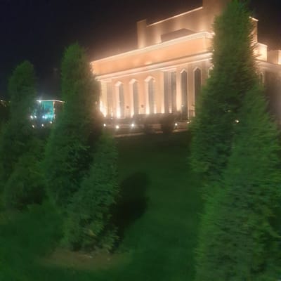 The image depicts a building at night, seemingly located in Turkistan, Kazakhstan, due to architectural styles common in that region. The building features a peach-colored facade with prominent columns, likely constructed from a modern material. The lights illuminate the structure, highlighting its architectural details. The building is surrounded by dark green vegetation, including trees and shrubs. The location is a garden.