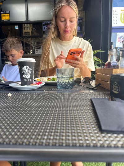A young woman with long blonde hair is holding an orange smartphone and looking down. She is wearing a light yellow t-shirt. Next to her is a glass of water with ice and a sprig of mint, and a small salad with a red tomato and what appears to be a piece of cheese. In the foreground, a young boy with short brown hair is wearing a white t-shirt and looking down at a black "TO GO" coffee cup with a decorative white design. The scene appears to be outdoors at a cafe or restaurant, with a textured dark gray table and a glimpse of green grass in the background. A wooden crate with bottles is visible behind the woman. The time of day appears to be daytime, with bright lighting suggesting good weather. The text visible includes "TO GO" on the coffee cup, and a label on a box or dispenser in the background that reads "Movistar". The woman's expression is calm, and the overall atmosphere seems relaxed.