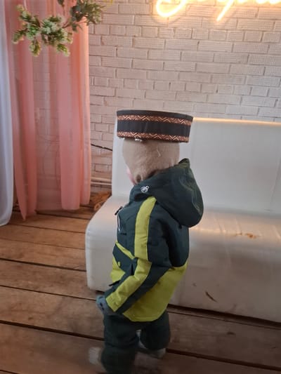 A vertical shot captures a child-sized mannequin or figure, oriented sideways in the provided image. The figure is dressed in a dark green and lime green winter jacket or snowsuit and wears a distinctive hat with geometric patterns in black, brown, and possibly white. A light-colored fabric is visible around its neck. The figure stands on a white, low platform or stool.
The setting appears to be an indoor space, likely a room or display area with a rustic aesthetic. On the left side of the frame, a glowing, bright light source (possibly a neon sign) illuminates a white textured wall resembling brickwork. Below the light, sheer pink curtains or fabric are draped. A small plant with white flowers or buds is partially visible in the lower-left foreground, in front of the curtains.
The floor is made of distinct wooden planks, extending across the bottom and to the right. Behind the figure, a plain white surface is visible, showing some reddish-brown stains or marks in the upper right portion. Further to the right, a vertical wooden structure or panel is present. No activity is taking place; the scene depicts a static arrangement. The time of day is not ascertainable, but the artificial light source suggests an indoor evening or controlled lighting environment. The location, Ludorvai, Russia, hints at a cold climate, which aligns with the winter clothing worn by the figure. There is no visible text.